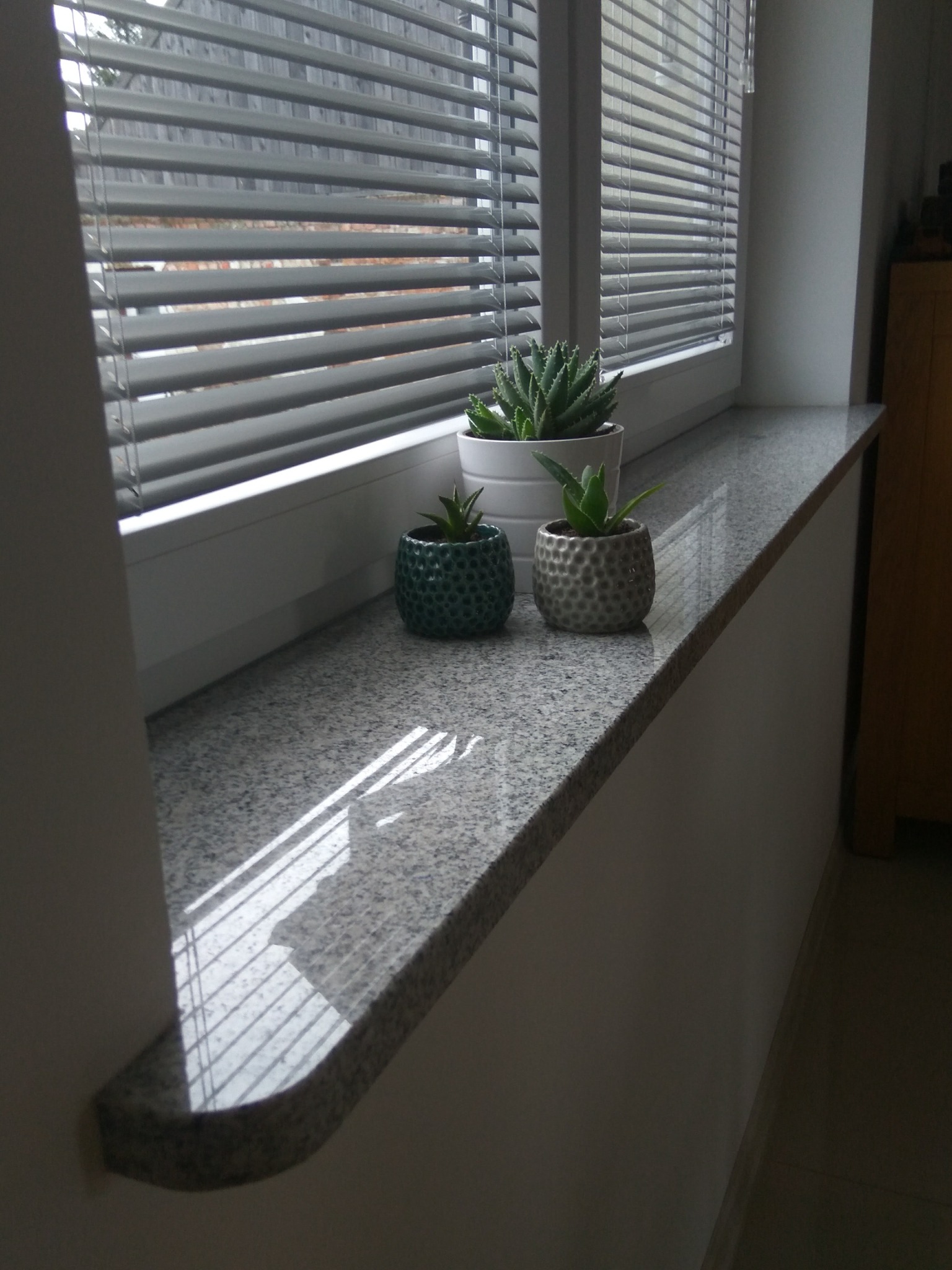 Szary, nakrapiany parapet z zaokrąglonym brzegiem, na którym stoją trzy małe doniczki z sukulentami; w tle okno z żaluzjami, przez które wpadają promienie słoneczne.