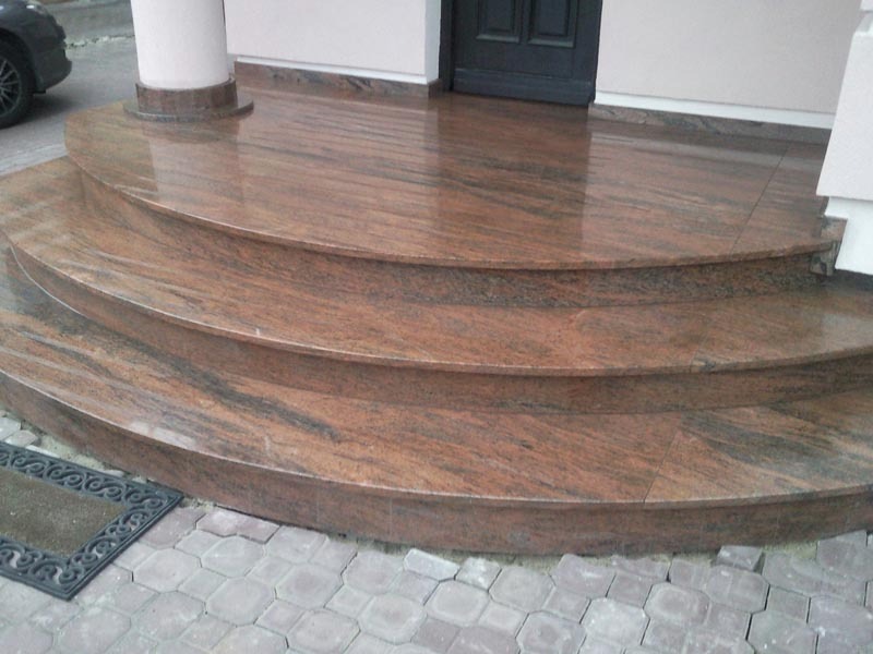 Zewnętrzne schody wejściowe wyłożone polerowanym kamieniem w odcieniach brązu i beżu, z widocznym wykończeniem cokołu filaru i fragmentem wycieraczki przy dolnym stopniu.