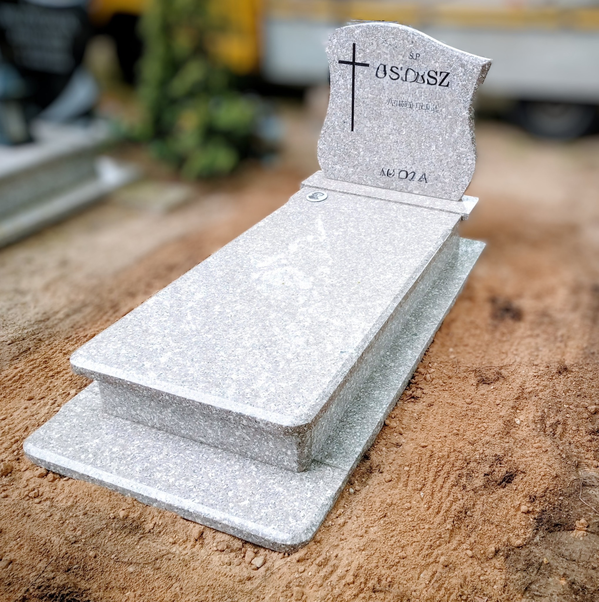 Jasnoszary, granitowy nagrobek z krzyżem, widoczny z perspektywy lekko z góry, na tle piaszczystej ziemi. Widoczny napis 'S.P. OS.D.SZ'.