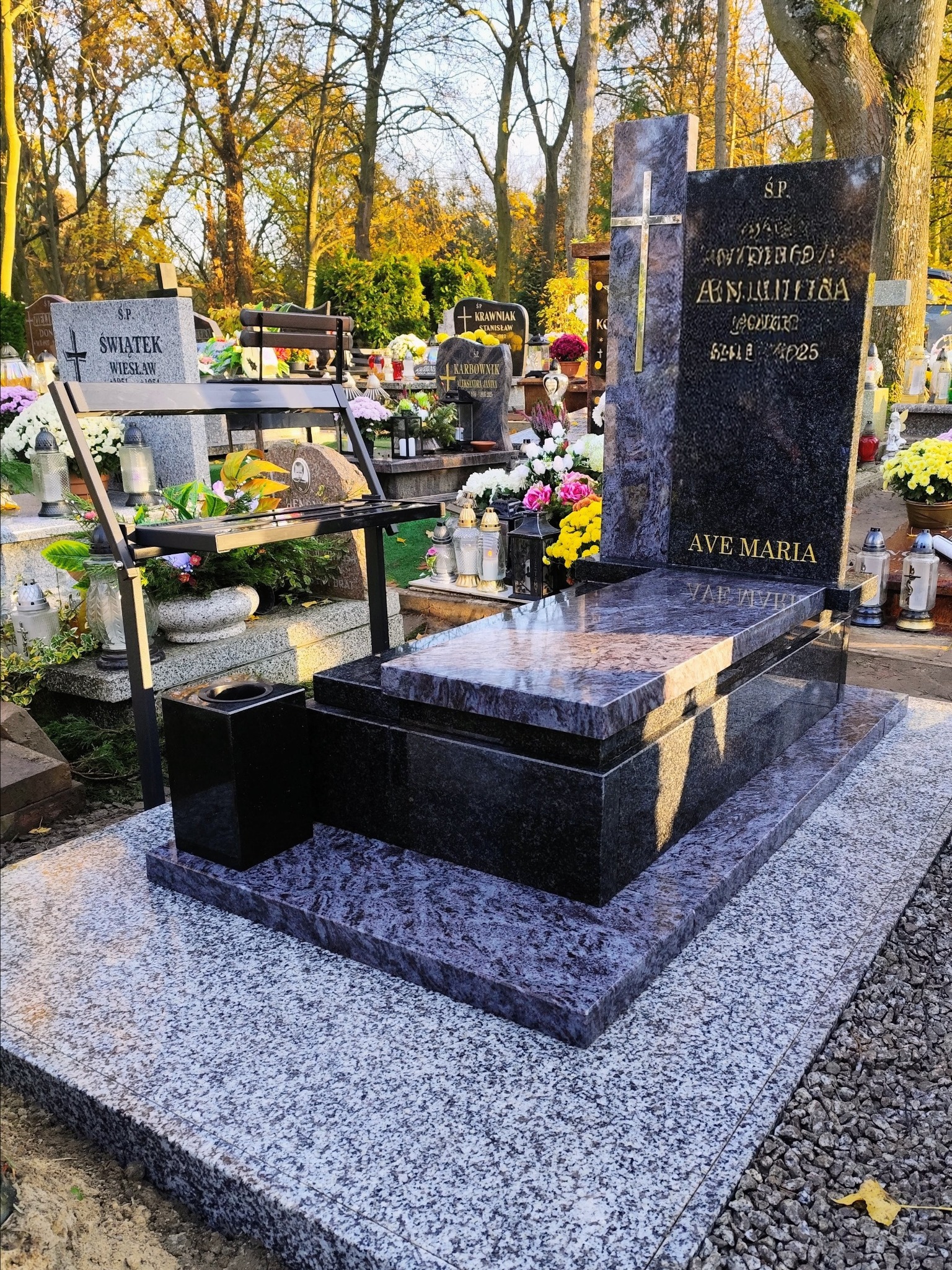 Granitowy nagrobek z krzyżem i napisem 'Ave Maria' na cmentarzu, otoczony zielenią i innymi nagrobkami. Widoczna ławka i kwiaty.