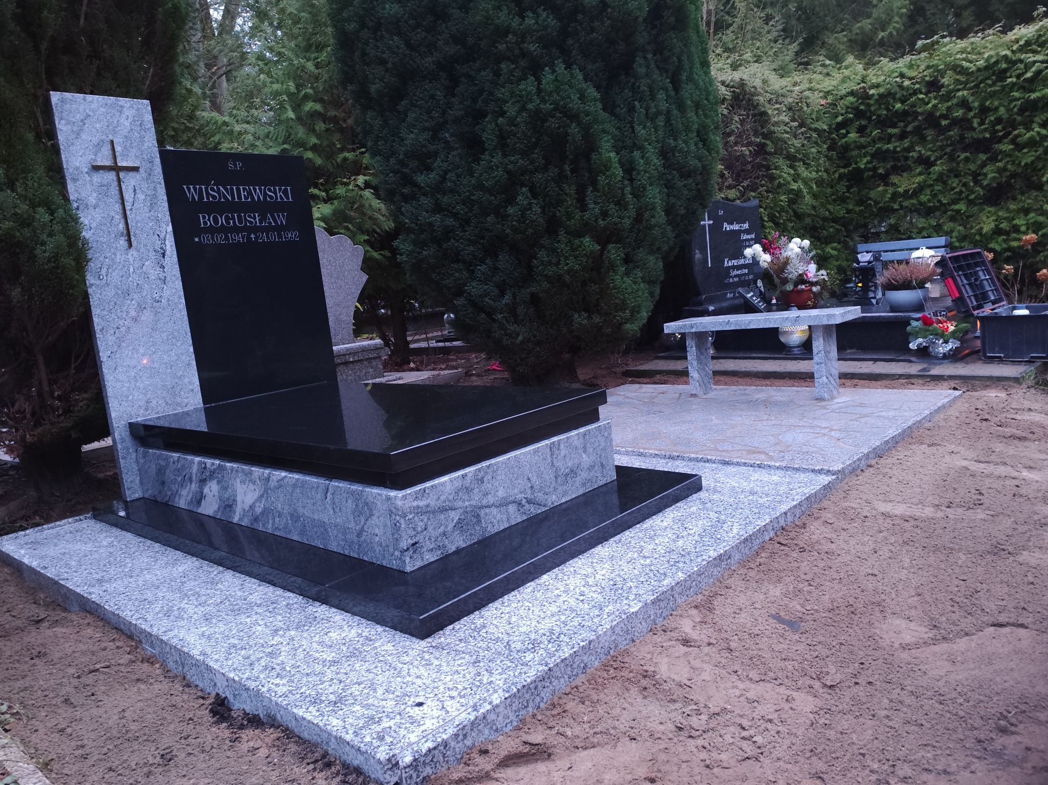 Granitowy nagrobek z czarną tablicą i krzyżem, otoczony zielenią cmentarza. Widoczne inskrypcje i data. Jasnoszara podstawa nagrobka.