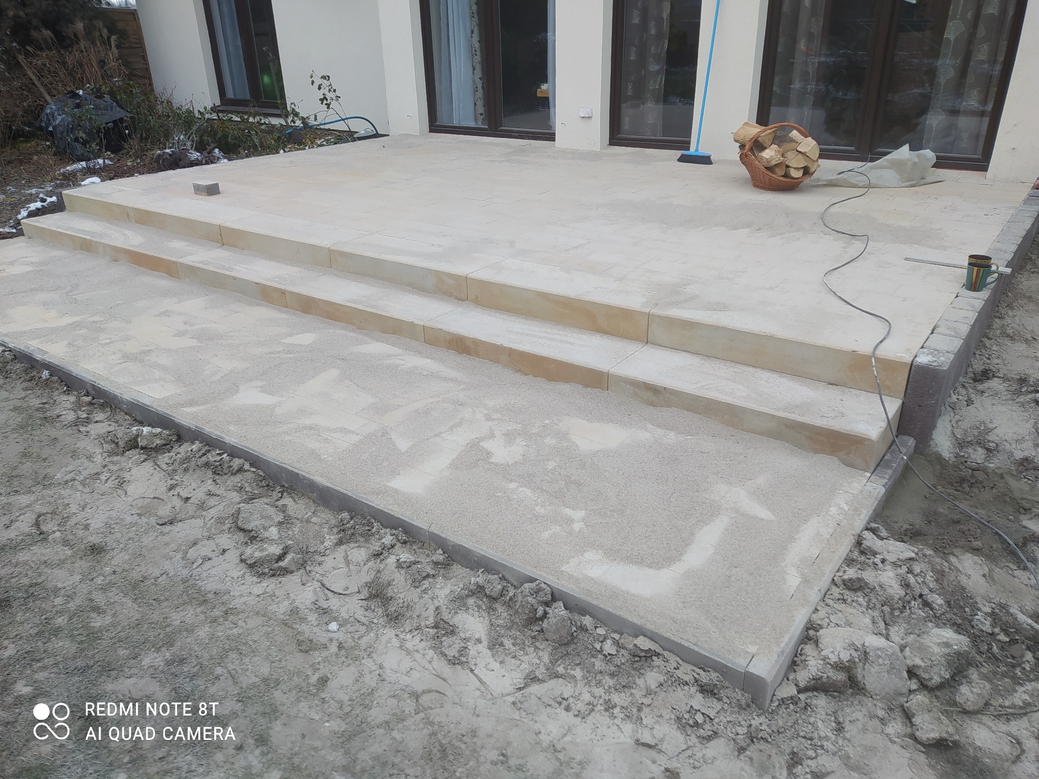 Jasny taras z kamiennymi stopniami w trakcie budowy, widoczne narzędzia i materiały budowlane, otoczenie w stanie surowym.
