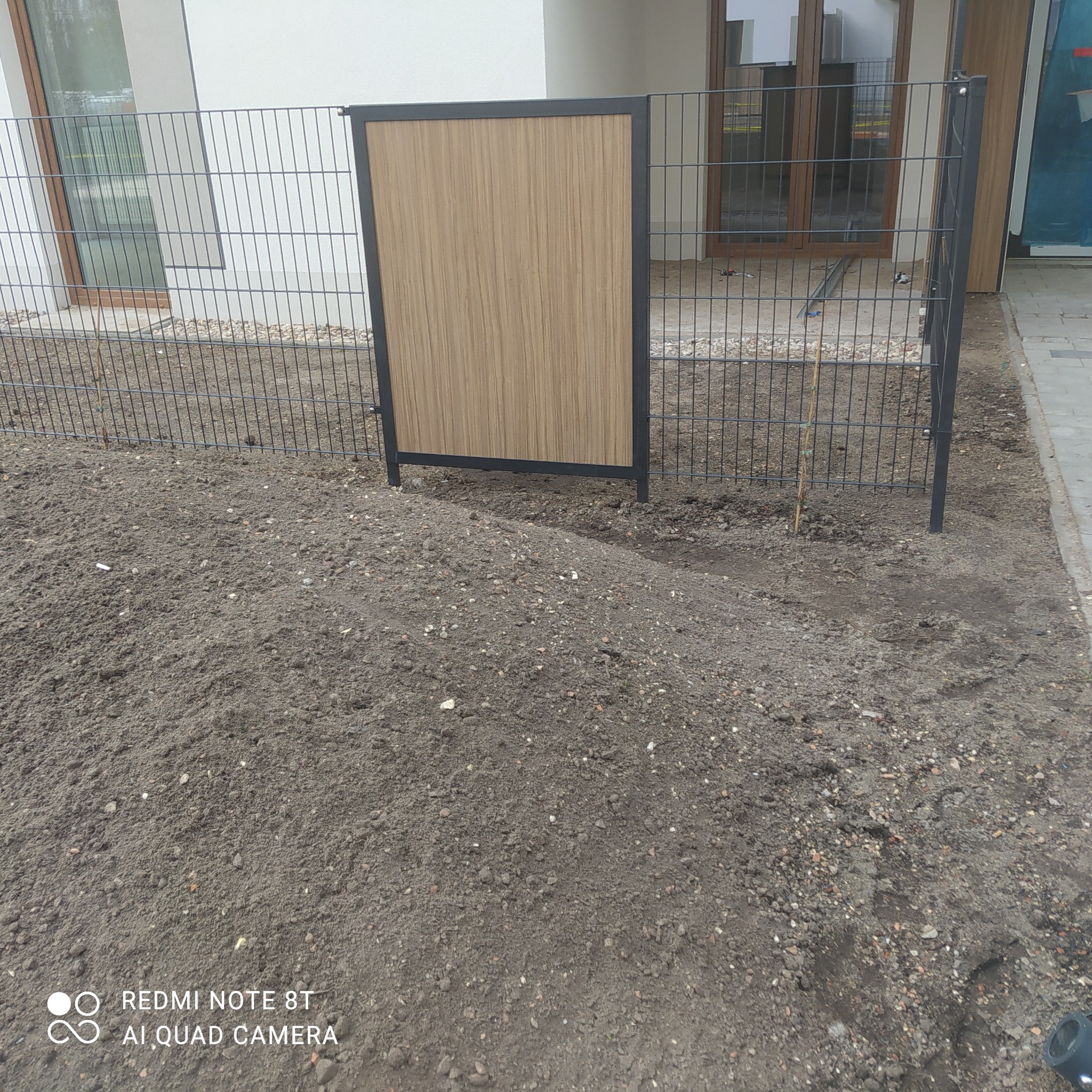 Montaż ogrodzenia panelowego z wypełnieniem imitującym drewno, widoczna świeżo usypana ziemia.