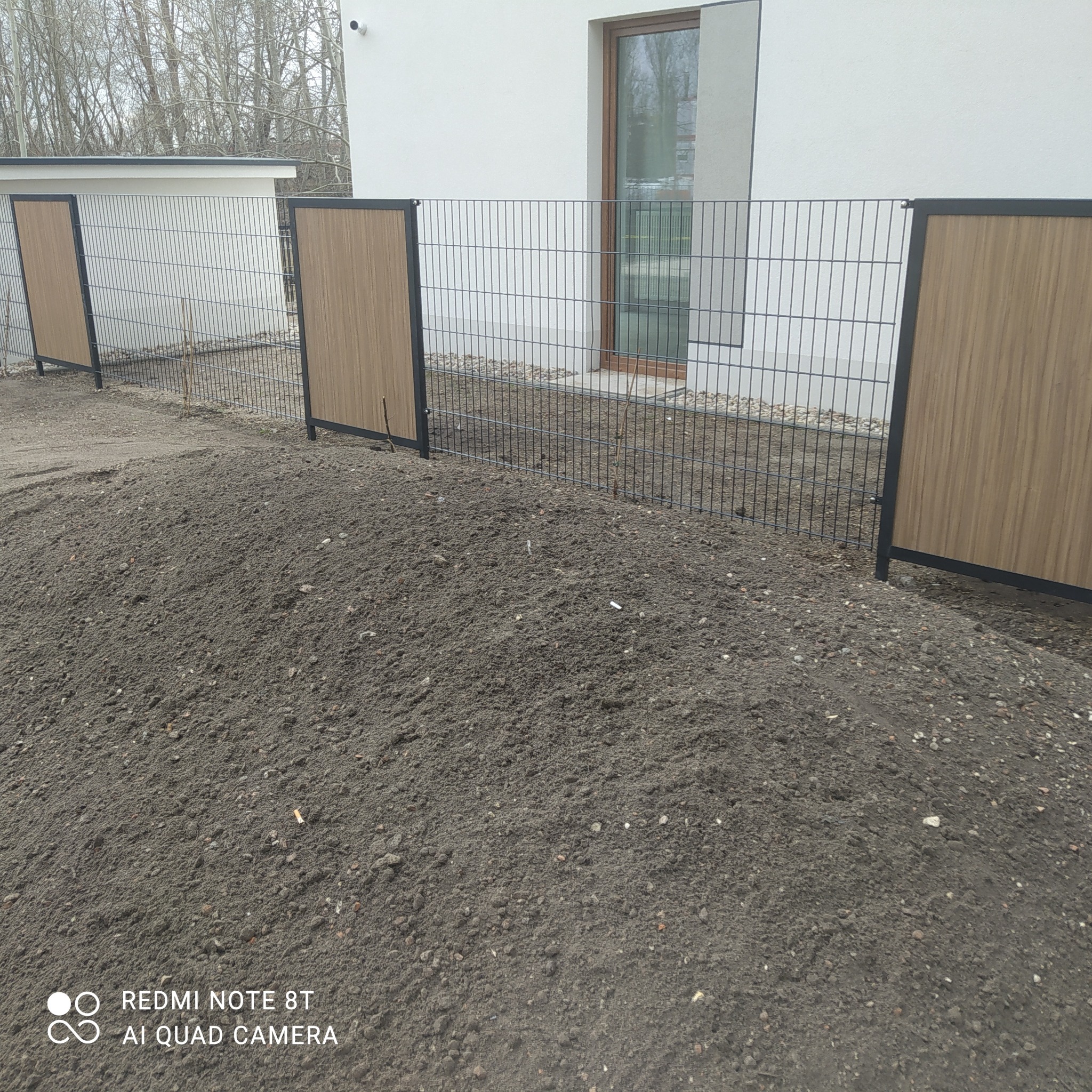 Nowoczesne ogrodzenie panelowe z elementami imitującymi drewno, widoczne na tle nowo usypanej ziemi przy budynku.