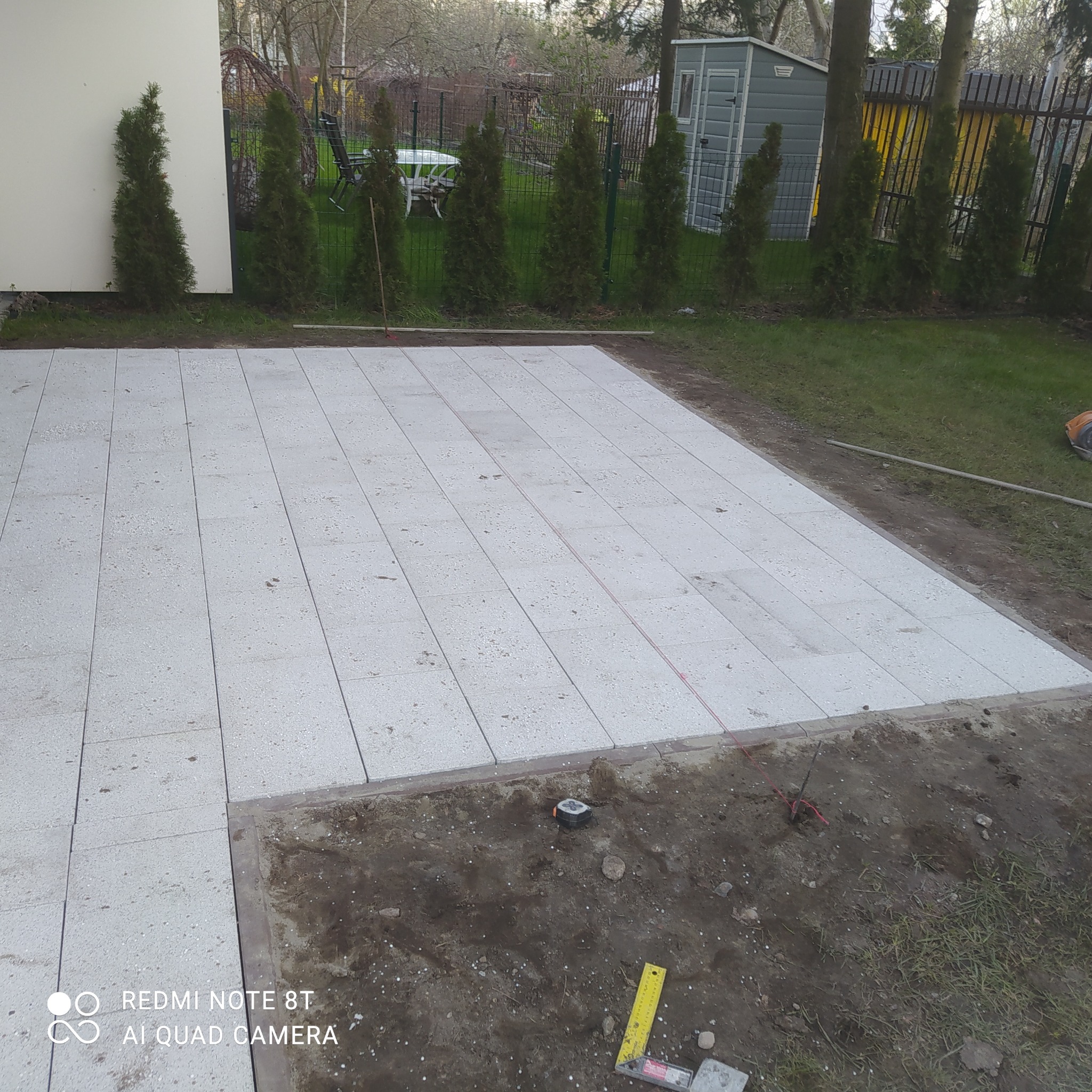 W trakcie budowy tarasu: ułożone płyty betonowe na przygotowanym podłożu, widoczne narzędzia miernicze i wytyczone sznurki, w tle rząd tuj i ogrodzenie.