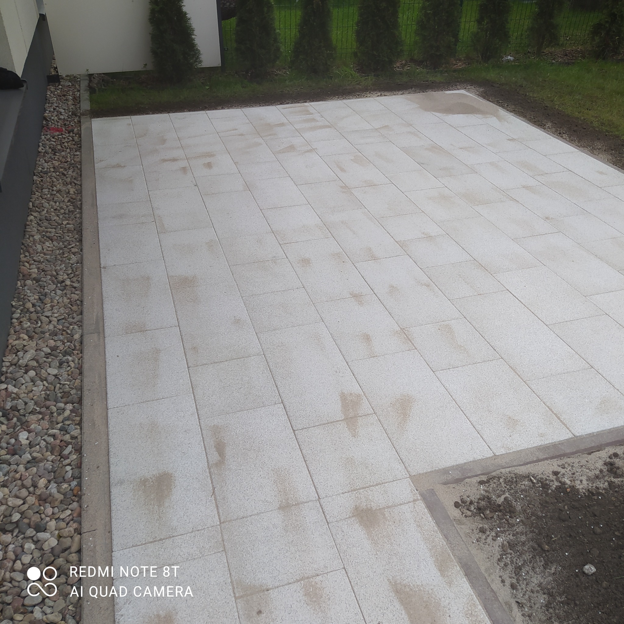 Jasnoszary taras z płyt betonowych ułożonych regularnie, obrzeże z drobnych kamieni przy ścianie budynku, w tle żywopłot z tuj.