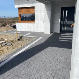 Nowoczesna posesja w trakcie wykańczania: ułożona szara kostka brukowa z białymi wstawkami prowadząca do drzwi wejściowych, widoczne prace ziemne i materiały budowlane w tle.