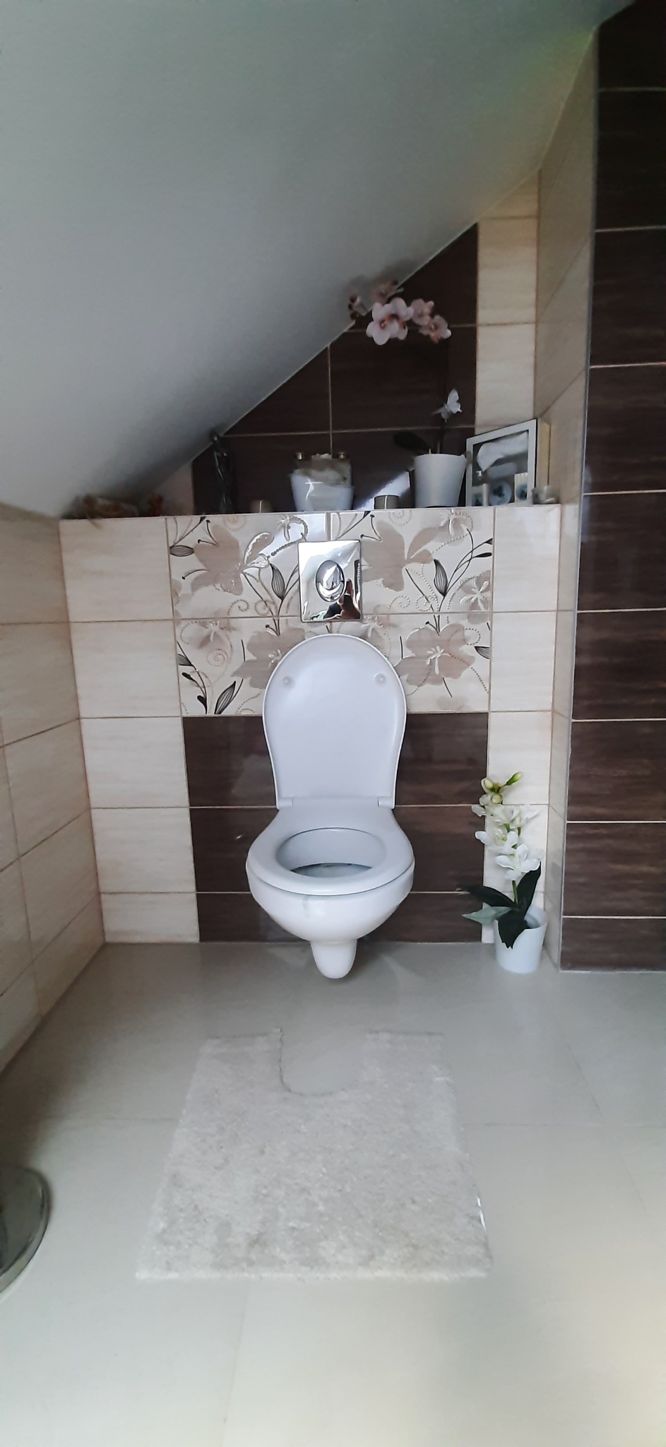 Kompaktowa toaleta podwieszana z chromowanym przyciskiem spłukującym, umieszczona w łazience z beżowo-brązowymi płytkami i dekoracyjnymi kwiatami.