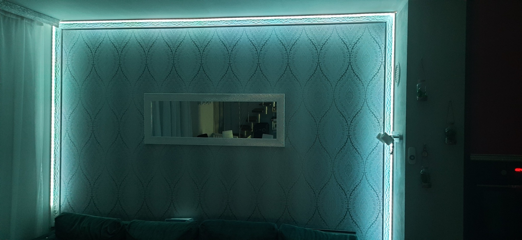 Ściana z wzorzystą tapetą oświetloną listwą LED, z zawieszonym lustrem w białej ramie.