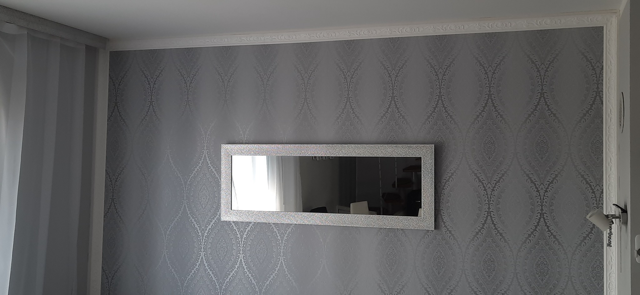 Ściana w srebrnej tapecie z ornamentem, ozdobna listwa przysufitowa i prostokątne lustro w ramie z mozaiki.