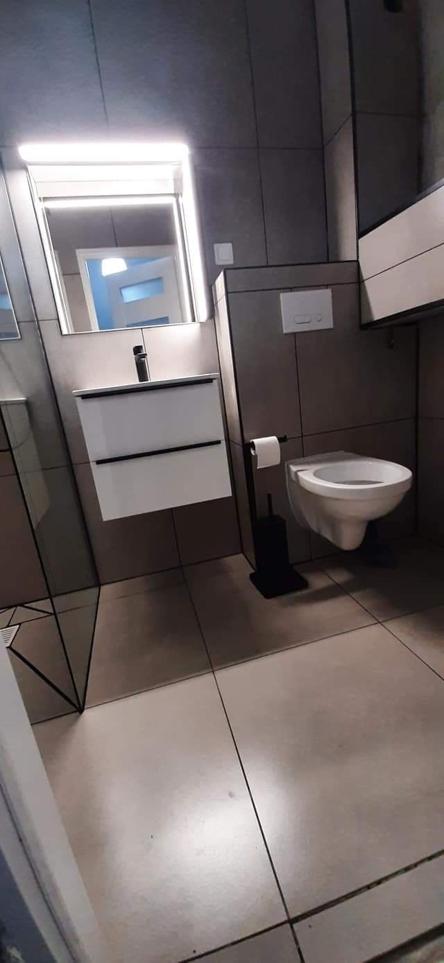 Nowoczesna łazienka z szarymi płytkami, podwieszaną toaletą i umywalką z szafką podświetloną ledowym lustrem. Widoczny dozownik mydła i uchwyt na papier toaletowy.