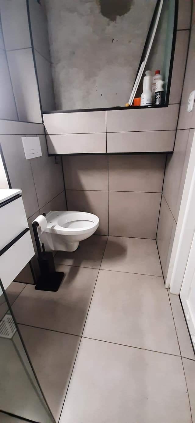 Wnętrze łazienki w trakcie wykańczania, widoczne szare płytki, podwieszana toaleta i szafka nad nią, ściana nad szafką nieotynkowana, butelki z chemią na szafce.