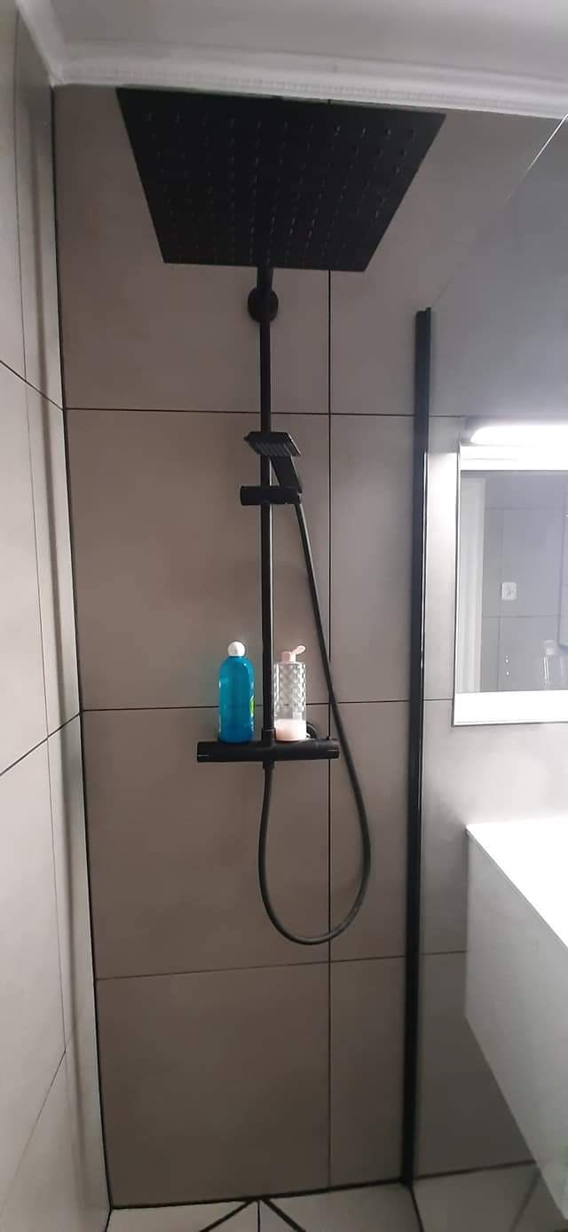 Nowoczesna kabina prysznicowa z czarną armaturą, szare płytki ceramiczne, półka z kosmetykami, widoczne odbicie lustra.