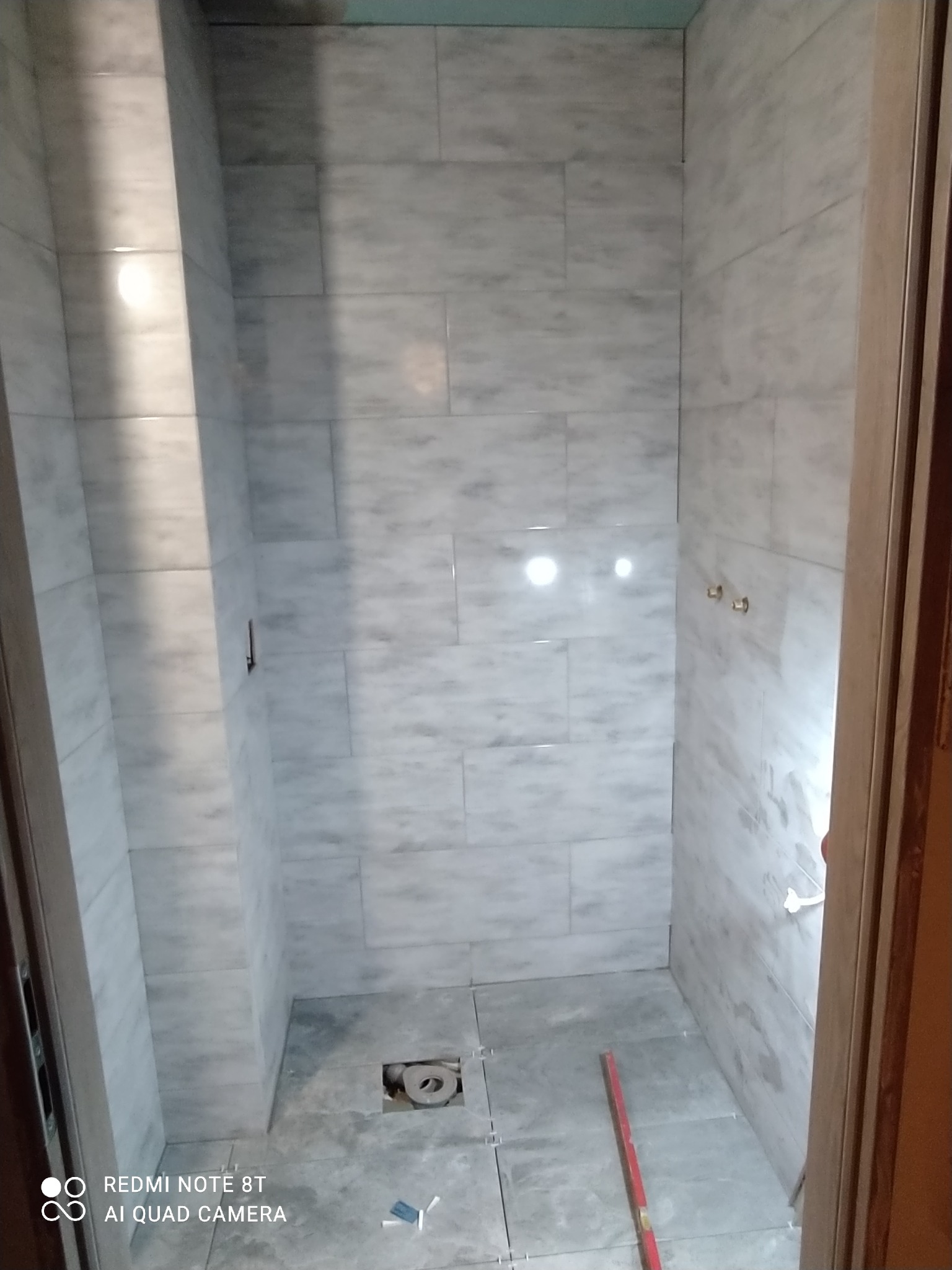 Wnętrze łazienki w trakcie układania płytek imitujących marmur, widoczne poziomica, krzyżyki dystansowe i otwór odpływowy.