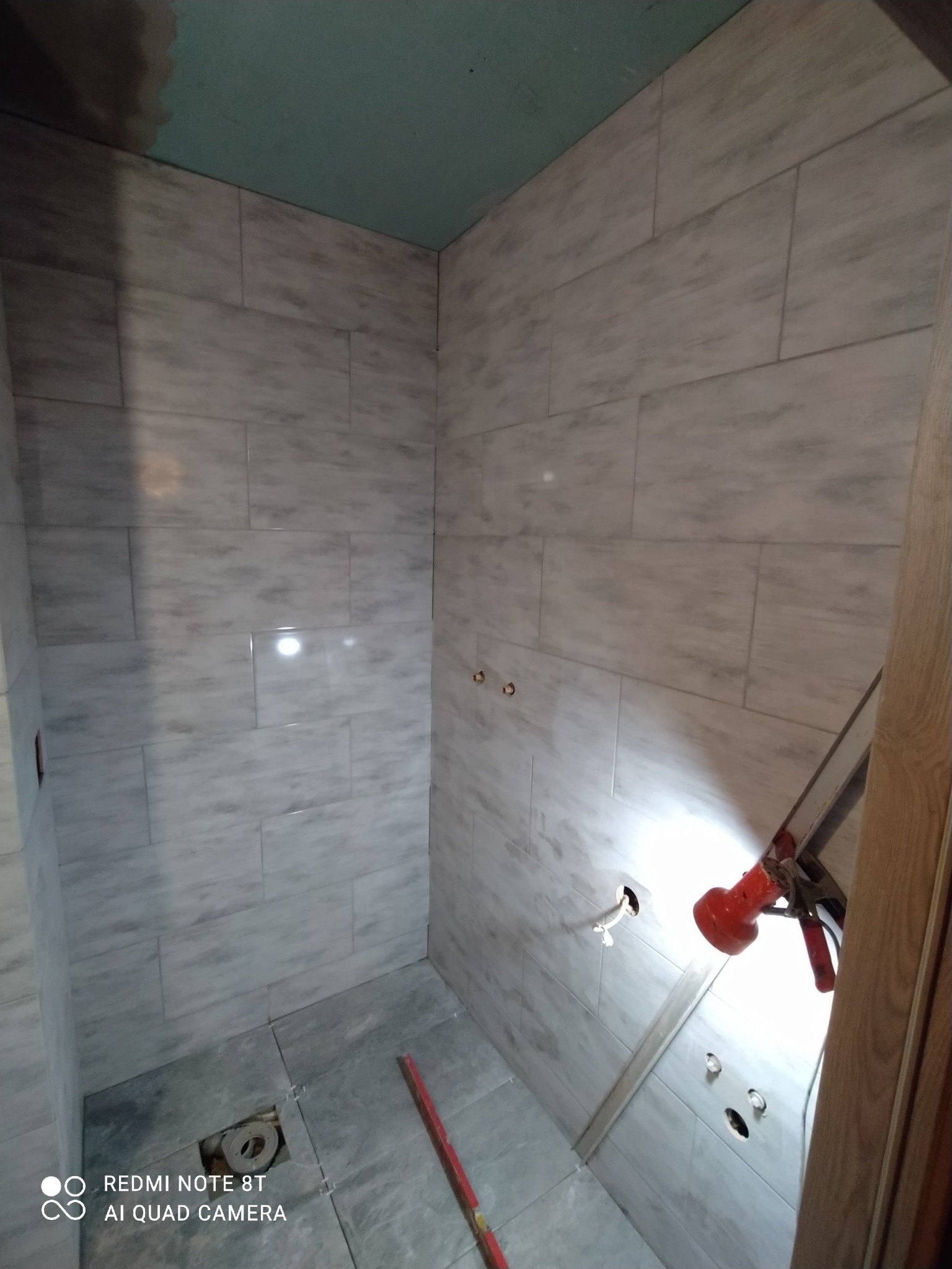 Wnętrze łazienki w trakcie układania płytek imitujących marmur, widoczne niedokończone fugi, rurki i narzędzia glazurnicze.