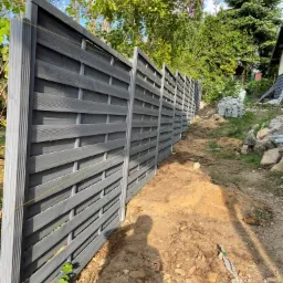 Długi, szary, drewniany płot panelowy w trakcie budowy, z widoczną linią poziomowania i stertą kostki brukowej w tle, otoczony zielenią.