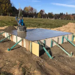 Świeżo wylany fundament z szalunkiem i pompą do osuszania terenu w tle, widoczne przygotowane pryzmy ziemi.