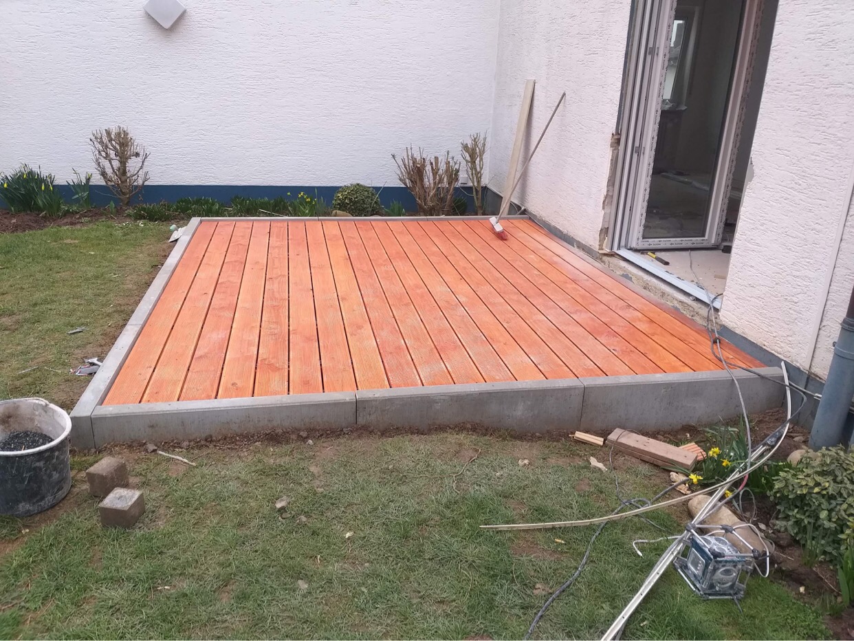 Nowo wybudowany taras z desek w kolorze pomarańczowym, obramowany betonową kostką, przylegający do białego budynku z otwartymi drzwiami balkonowymi, widoczne narzędzia i resztki materiałów...