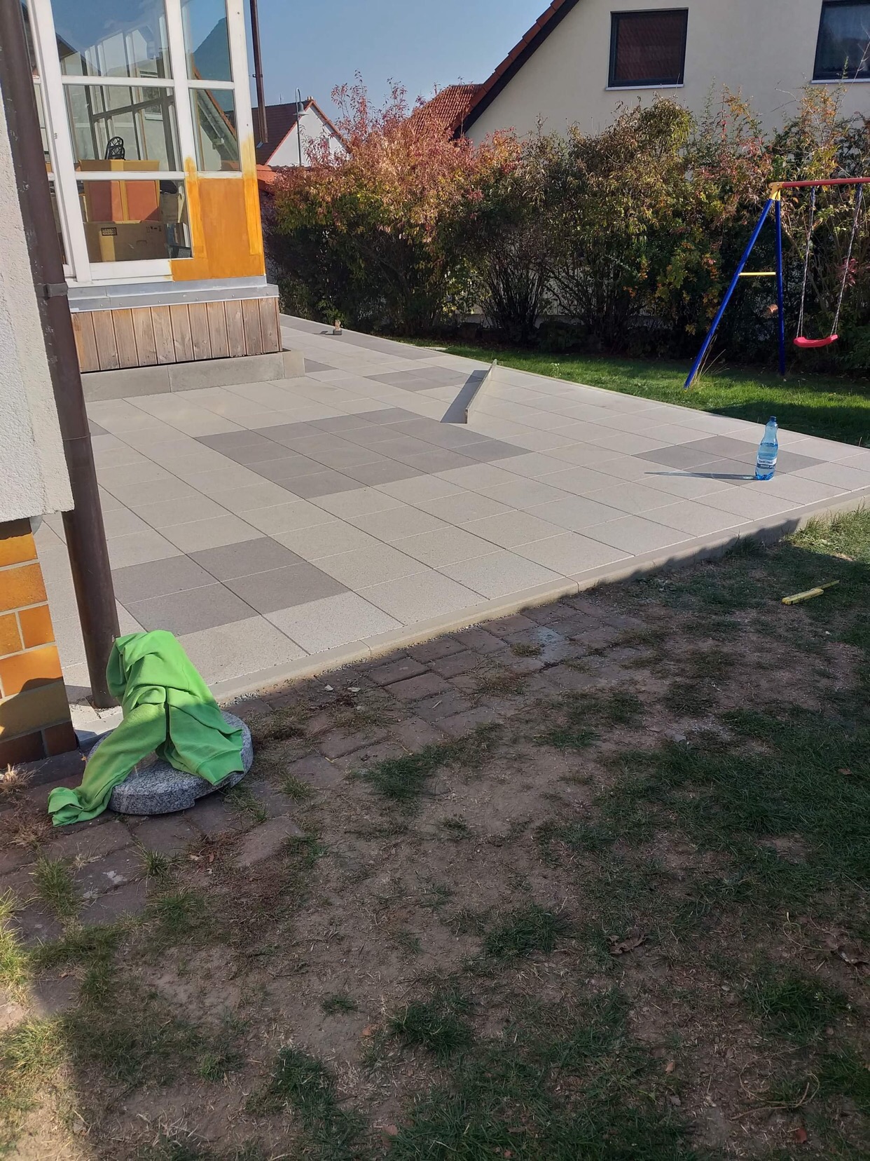 Nowa, szara nawierzchnia z kostki brukowej ułożona przed domem z widoczną huśtawką w tle i butelką wody na obrzeżu, z zieloną szmatką leżącą na kamieniu przy ścianie budynku.