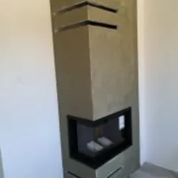 Narożny wkład kominkowy obudowany szarym, surowym betonem architektonicznym z poziomymi szczelinami wentylacyjnymi.