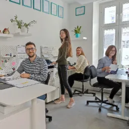 Wnętrze biura z czterema osobami przy biurkach, jedna osoba stoi przy kserokopiarce, na ścianach wiszą oprawione certyfikaty i półki z dekoracjami.