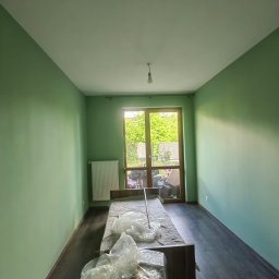 BestTiler - Pokój po świeżym malowaniu ścian na zielono, zabezpieczone meble folią, widoczne panele podłogowe i okno z widokiem na zieleń.
