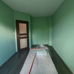 BestTiler - Pokój w odcieniach zieleni po malowaniu, zabezpieczone folią meble, widoczny wałek malarski z czerwoną rączką, ciemna podłoga, brązowe drzwi z przeszkleniami.