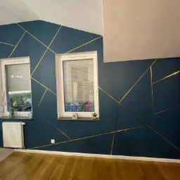 Ciemnoniebieska ściana z geometrycznym wzorem ze złotych linii, dwa okna z żaluzjami, drewniana podłoga, biały grzejnik i listwy przypodłogowe.