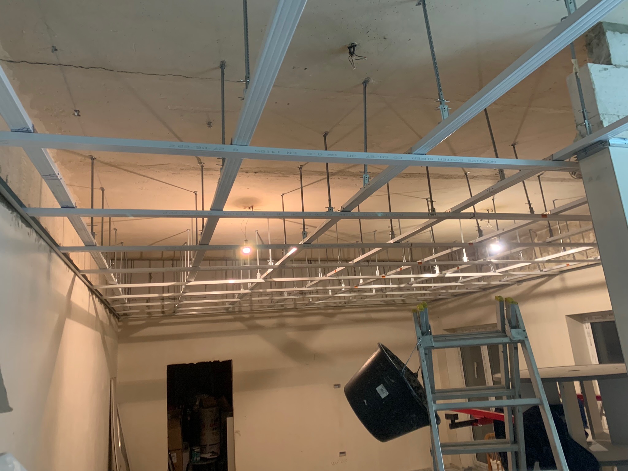 Metalowa konstrukcja sufitu podwieszanego z widocznymi profilami CD i UD, zawieszona na prętach mocujących, z surowym betonowym stropem w tle. Na pierwszym planie drabina aluminiowa i czarne wiadro...