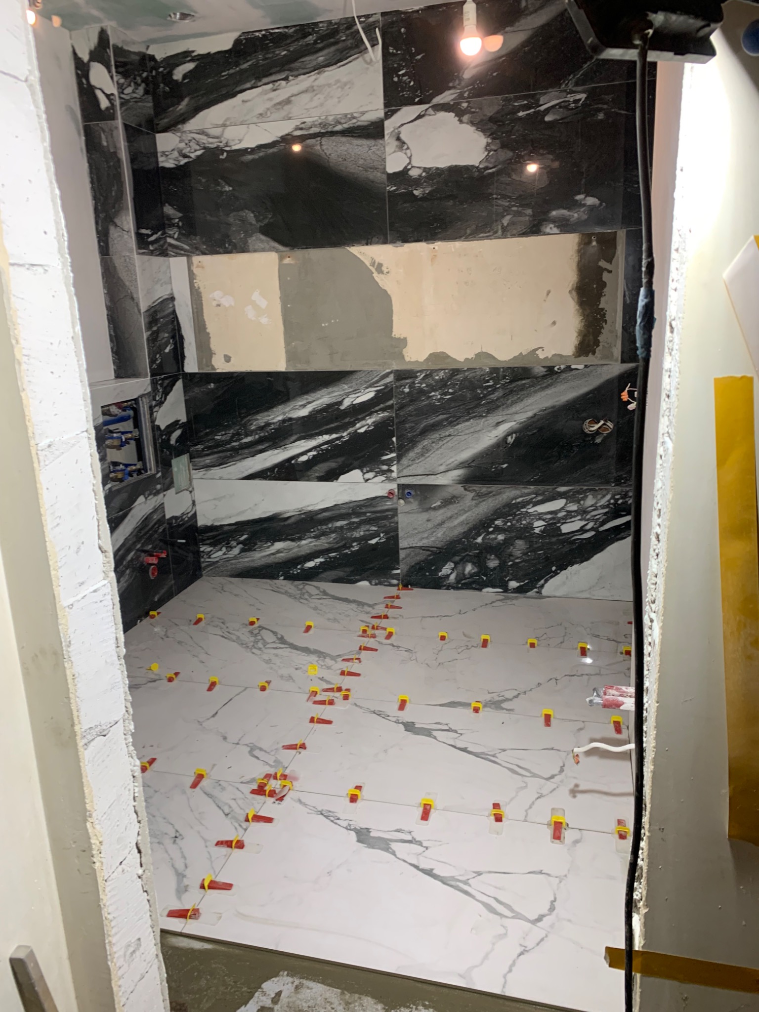 Łazienka w trakcie wykańczania: czarno-białe marmurowe płytki na ścianach i podłodze, z widocznymi elementami instalacji wodnej i poziomującymi klipsami do płytek na podłodze.