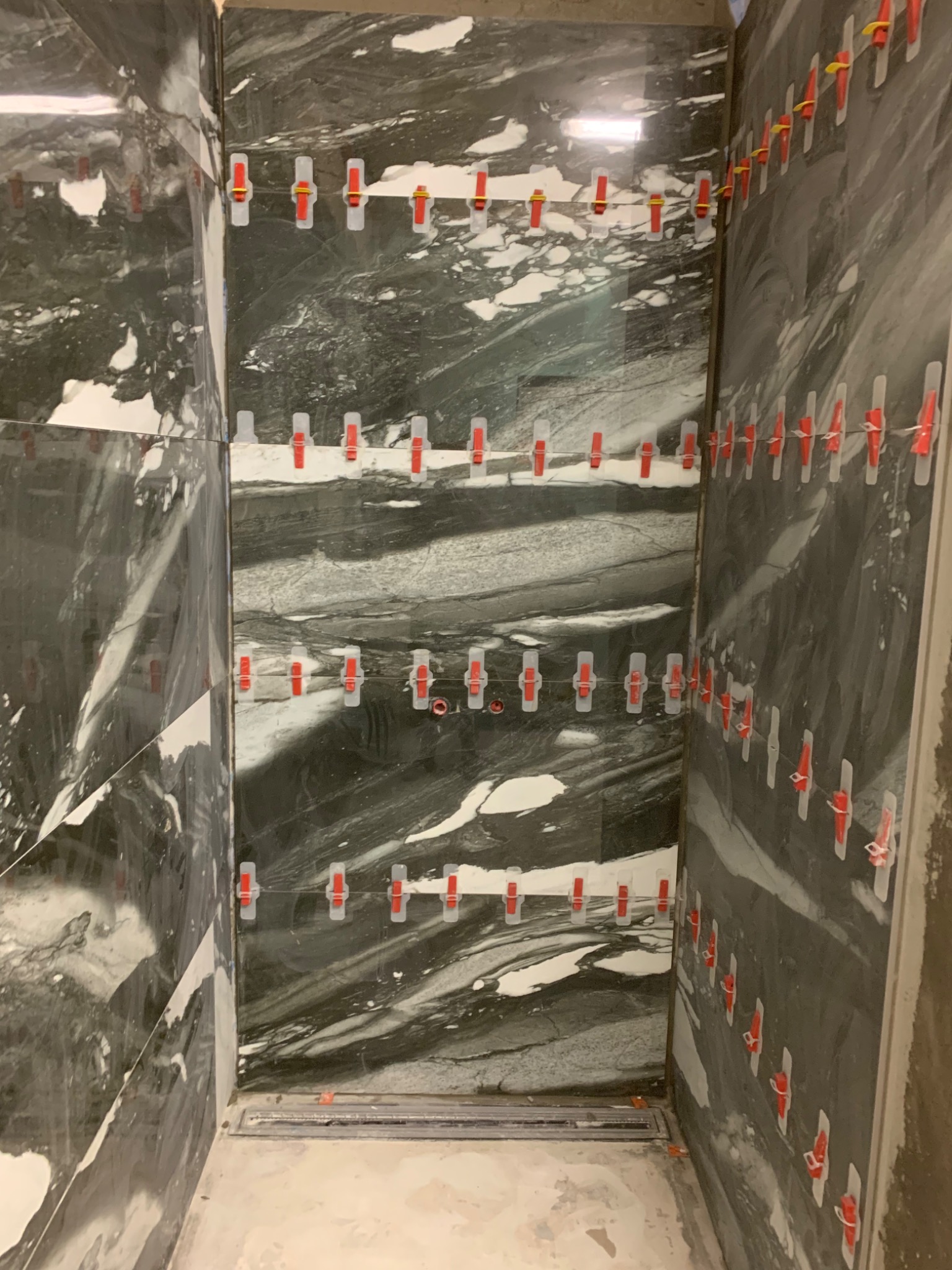 Układanie marmurowych płytek w kabinie prysznicowej z użyciem systemu poziomowania płytek z czerwonymi klipsami, widoczny odpływ liniowy.