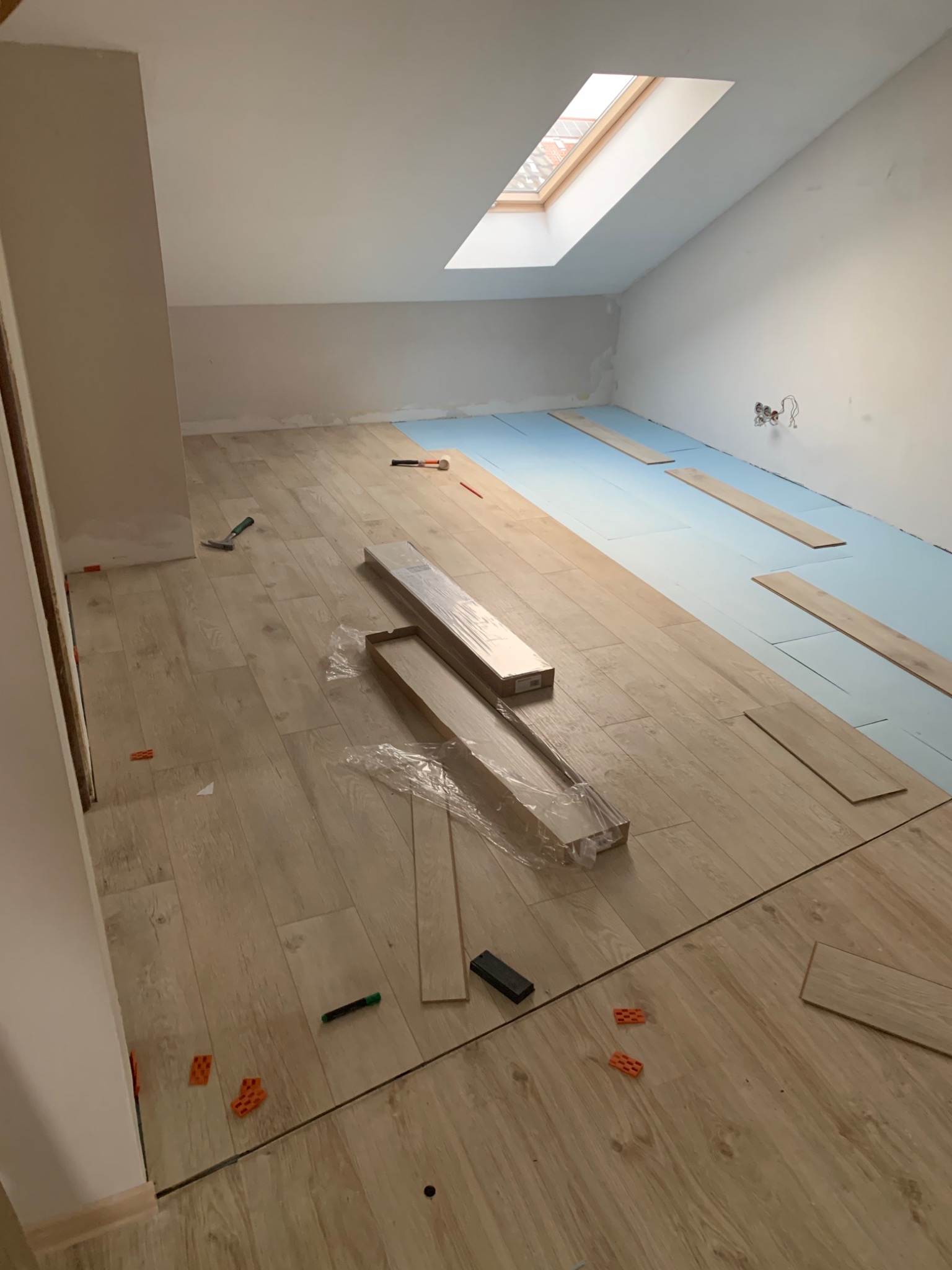 Układanie jasnych paneli podłogowych w pokoju na podkładzie izolacyjnym, widoczne narzędzia, kliny dystansowe i okno dachowe.