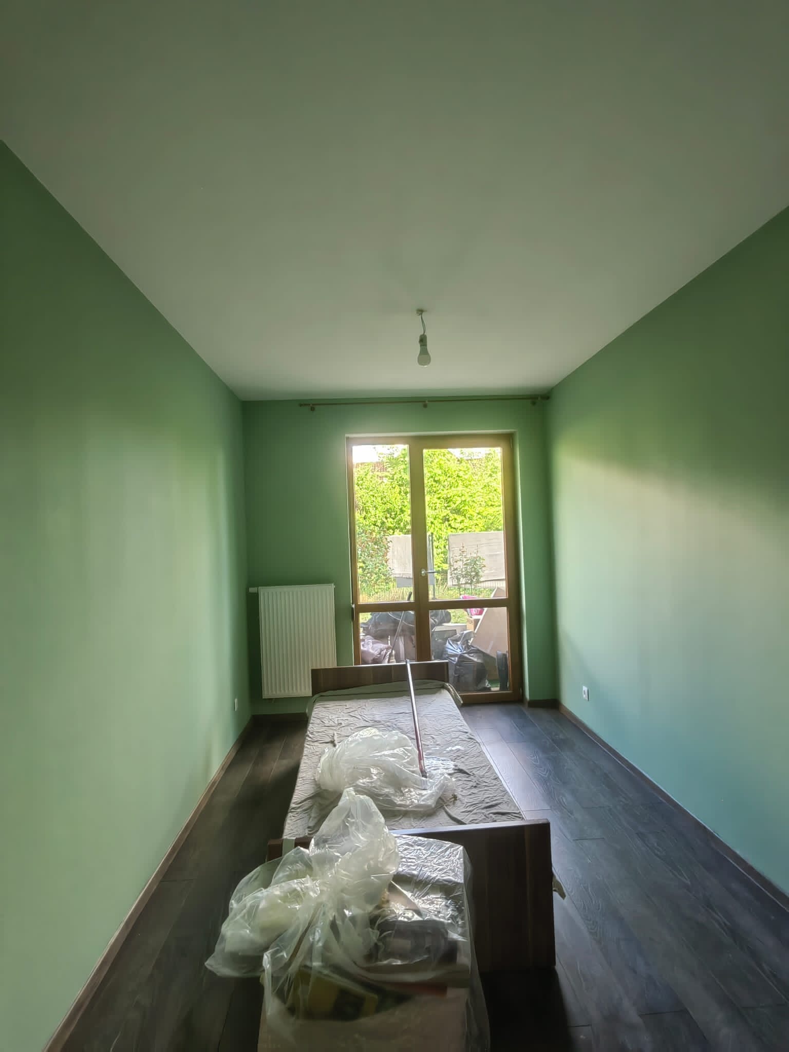 Pokój po świeżym malowaniu ścian na zielono, zabezpieczone meble folią, widoczne panele podłogowe i okno z widokiem na zieleń.