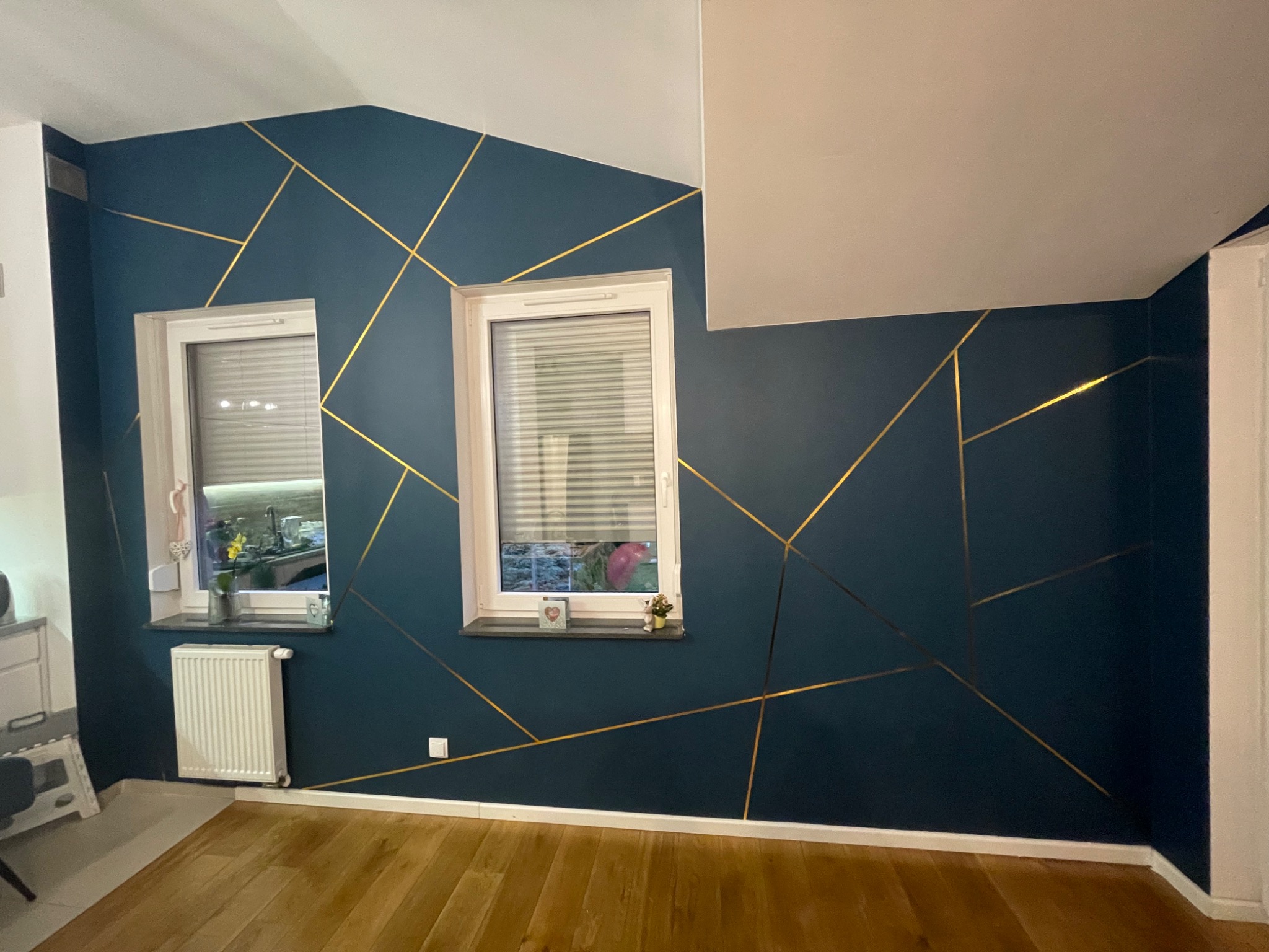 Ciemnoniebieska ściana z geometrycznym wzorem ze złotych linii, dwa okna z żaluzjami, drewniana podłoga, biały grzejnik i listwy przypodłogowe.