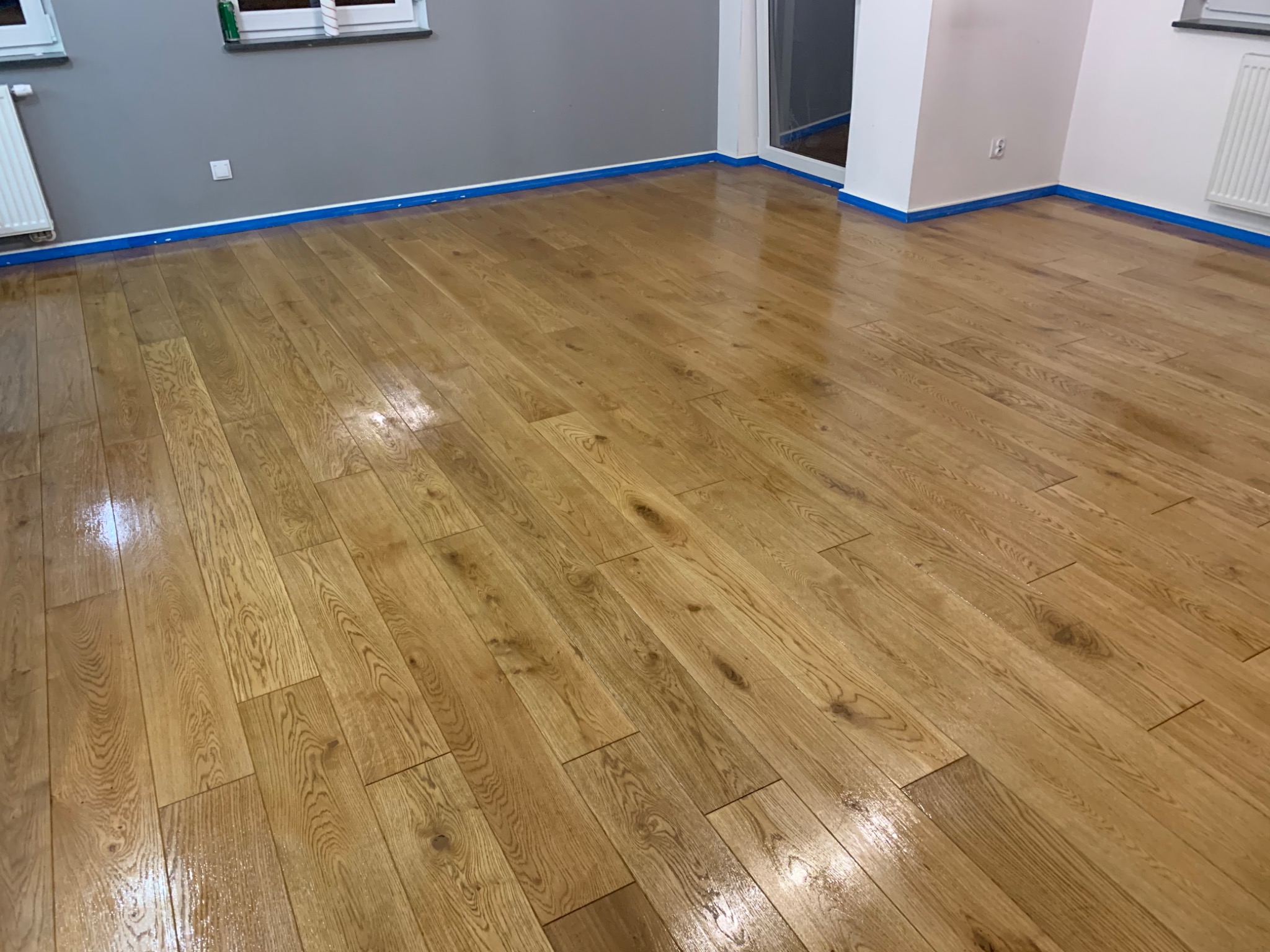 Świeżo położona, lakierowana podłoga parkietowa z wyraźnym usłojeniem, zabezpieczona niebieską taśmą malarską przy ścianach w szarym i białym kolorze.