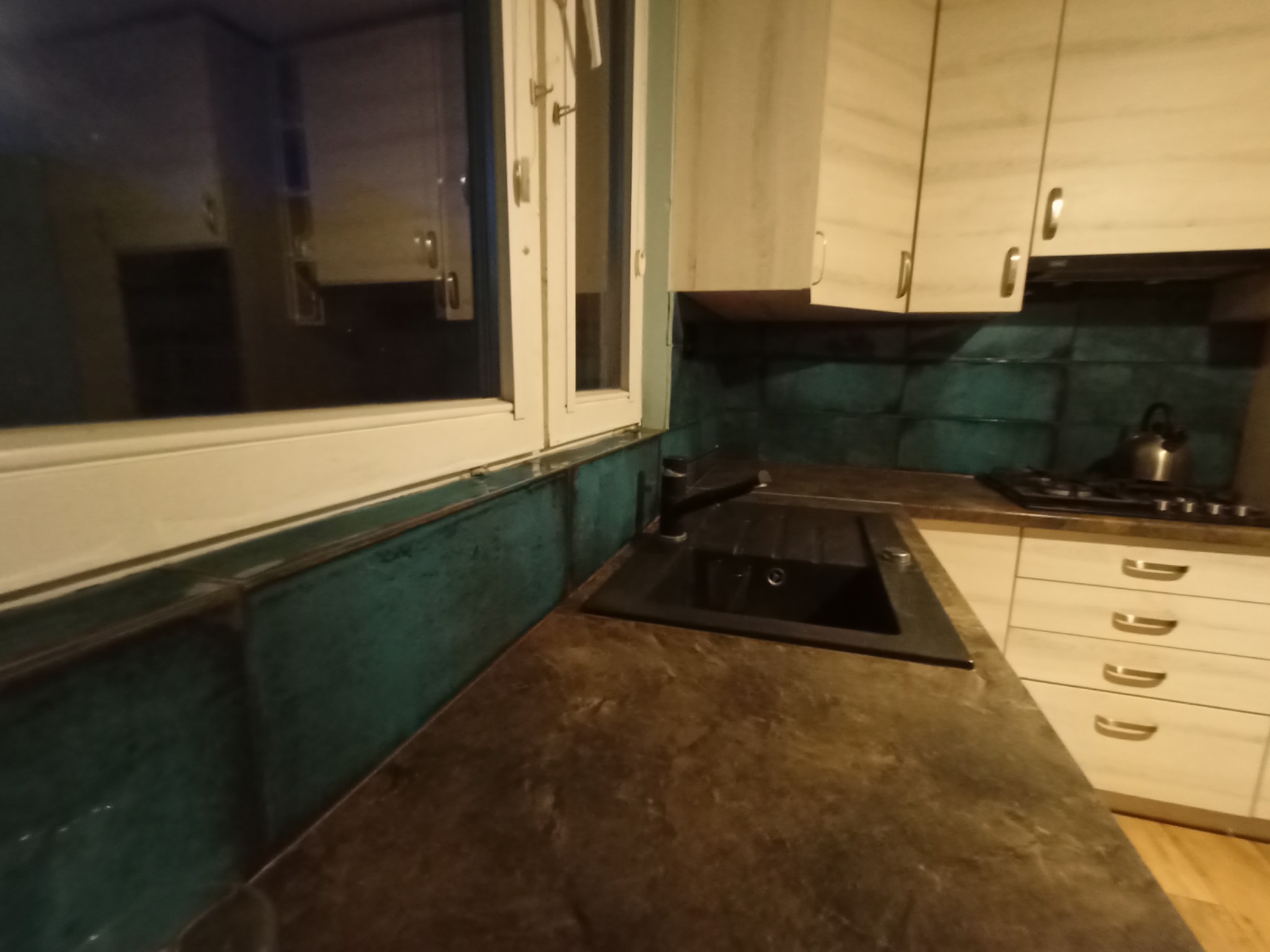 Kuchnia po remoncie: ciemnozielone płytki nad blatem, brązowy blat z czarnym zlewem, beżowe szafki i okno z białymi ramami.