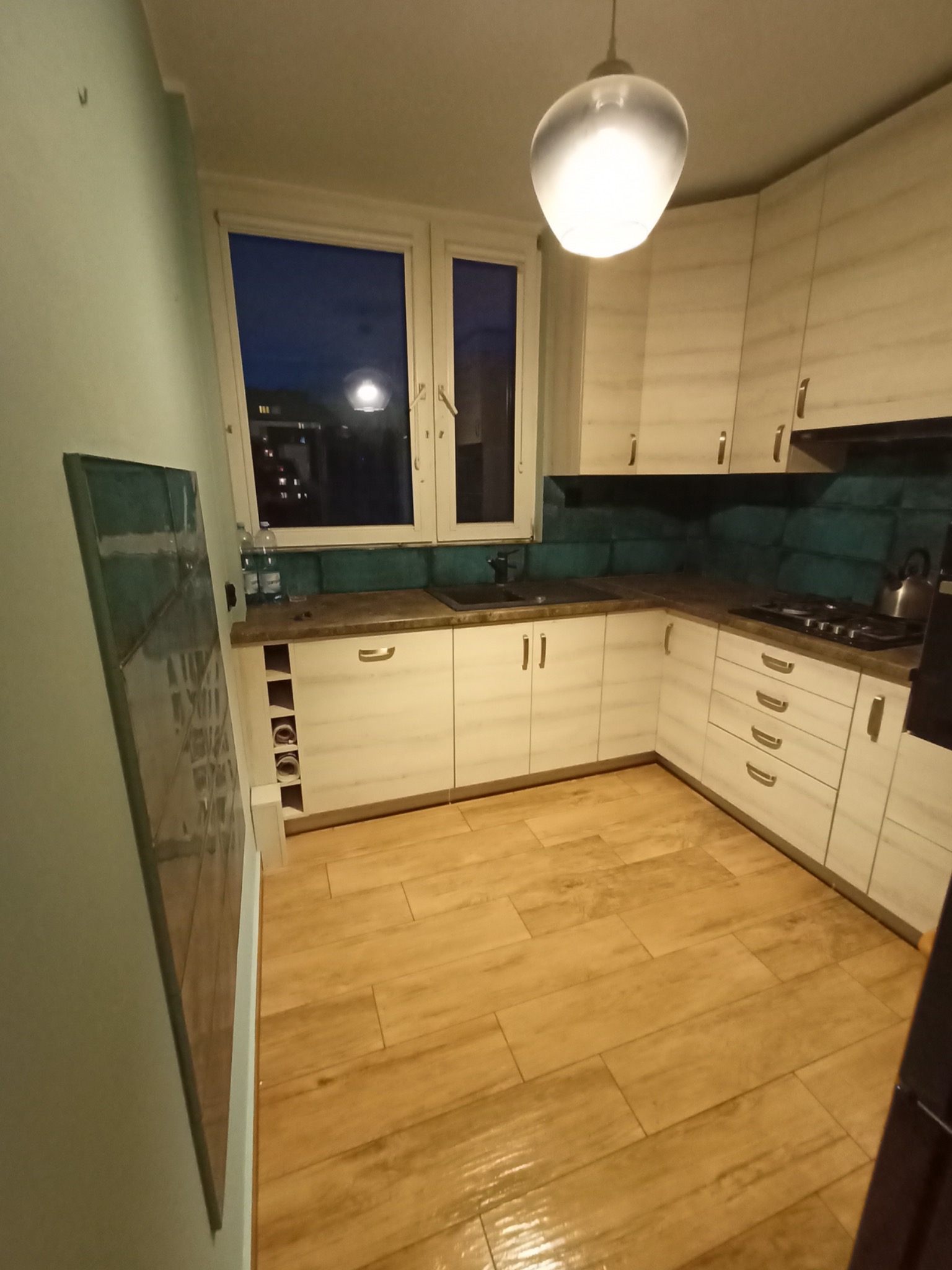 Ukończona kuchnia z beżowymi szafkami, turkusową glazurą nad blatem i drewnopodobną podłogą, widziana z niskiego kąta, z widokiem na nocne miasto przez okno.