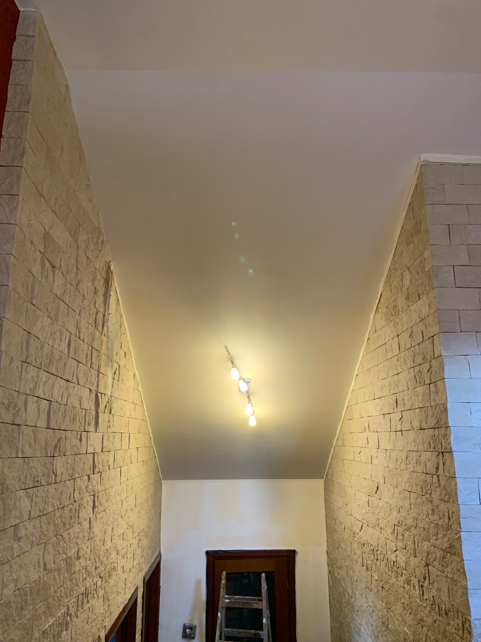 Wnętrze korytarza z dekoracyjną ścianą z kamienia, gładkim sufitem z oświetleniem punktowym i widoczną drabiną w tle.