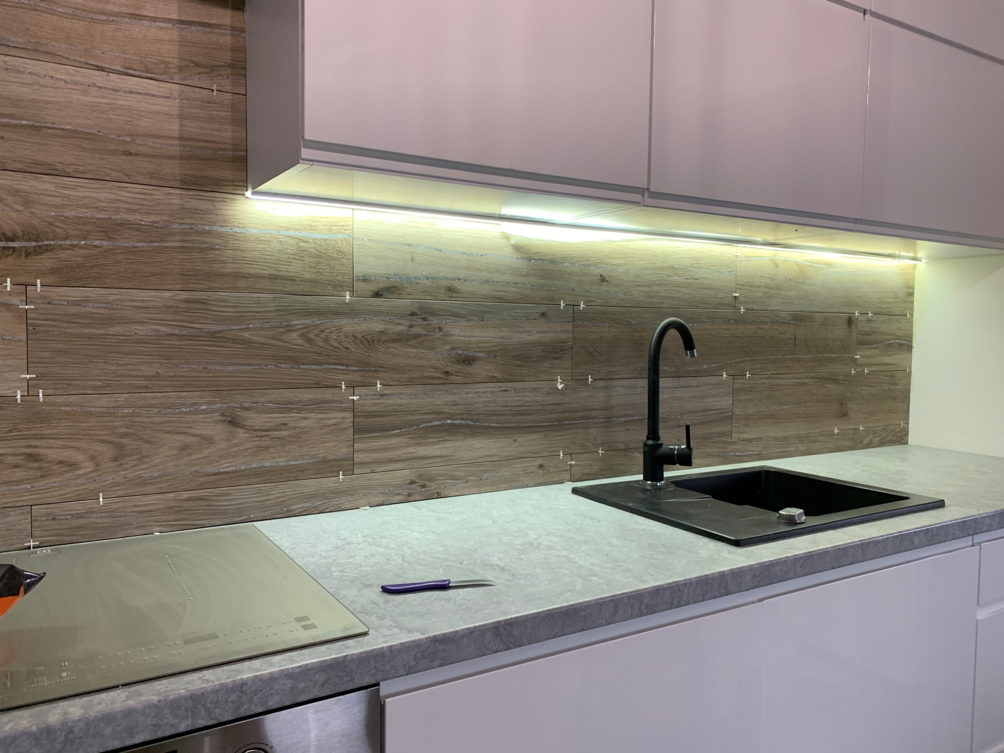 Kuchnia z drewnopodobnymi płytkami na ścianie, czarną baterią i zlewozmywakiem, płyta indukcyjna i białe szafki, widoczne krzyżyki dystansowe między płytkami, nóż na blacie.