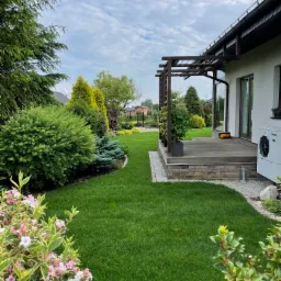 Zadbany trawnik z różnorodną roślinnością ozdobną, widoczny taras z drewnianą pergolą i jednostką zewnętrzną pompy ciepła przy nowoczesnym domu.