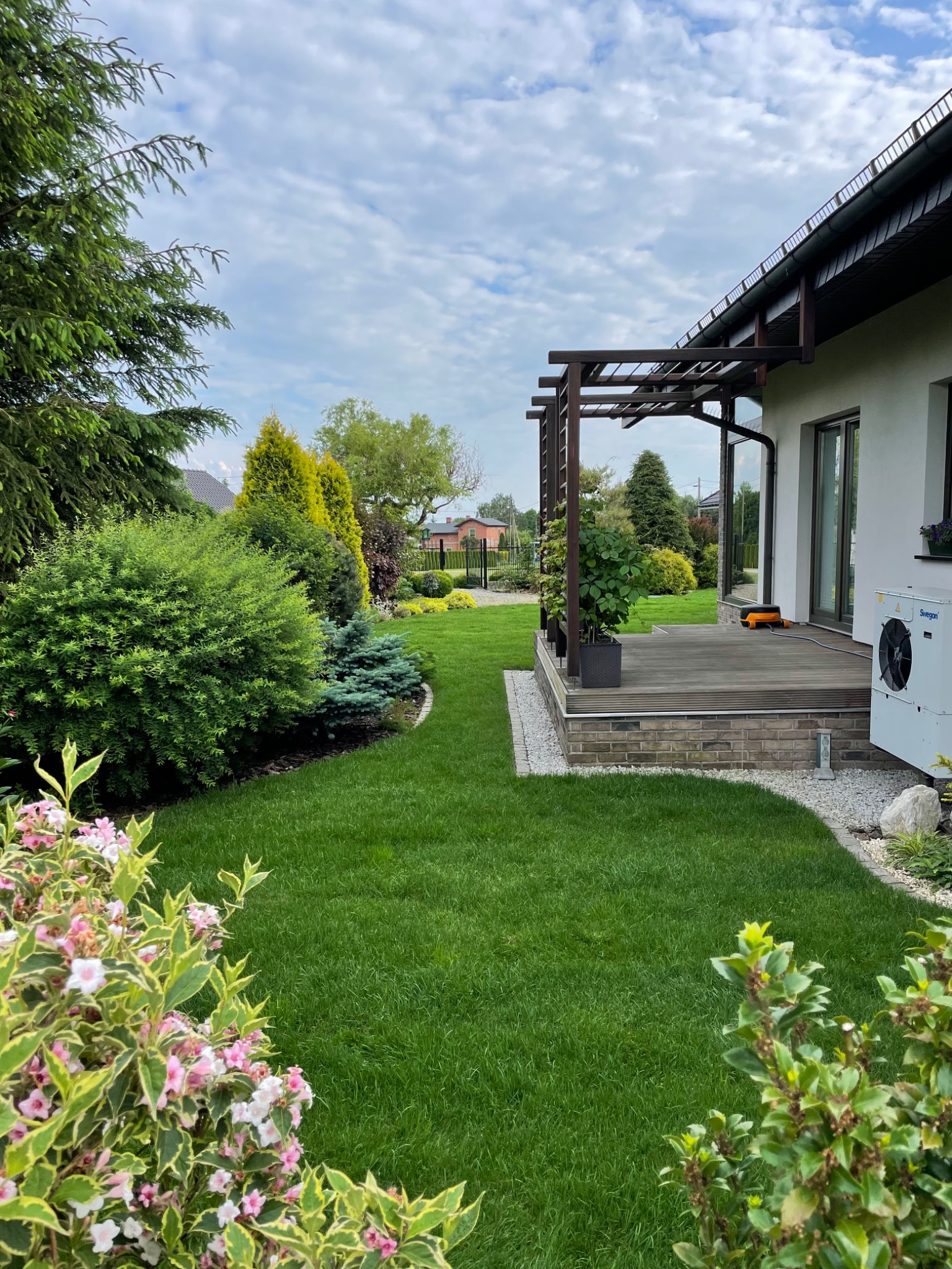 Zadbany trawnik z różnorodną roślinnością ozdobną, widoczny taras z drewnianą pergolą i jednostką zewnętrzną pompy ciepła przy nowoczesnym domu.