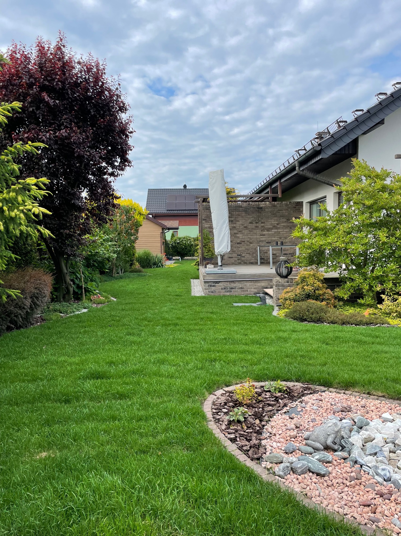 Zadbany trawnik z dekoracyjnym klombem z kamieni i figurką żaby, widok na taras z cegły i bujną roślinność w ogrodzie przydomowym.