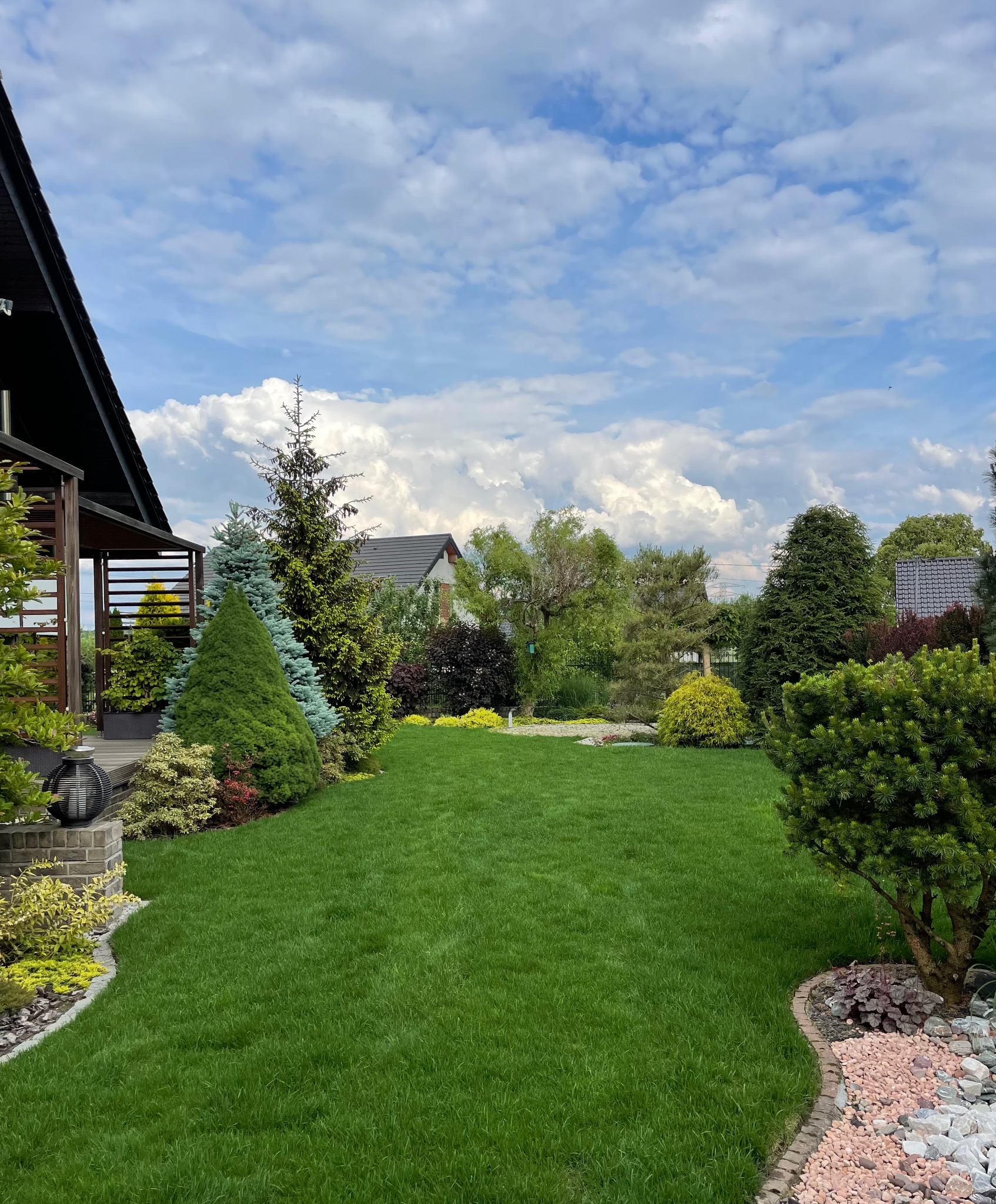 Zadbany trawnik z równo przystrzyżoną trawą, otoczony różnorodnymi drzewami iglastymi i krzewami ozdobnymi, z widocznym fragmentem domu z drewnianą pergolą i dekoracyjnym oświetleniem, na tle...