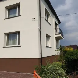 Dwukondygnacyjny dom z elewacją w kolorze złamanej bieli i brązowym cokołem, widoczny z perspektywy chodnika z pomarańczową barierką, z fragmentem balkonu i zieleni w tle.