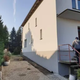 Dwukondygnacyjny budynek z ocieploną i pomalowaną na biało elewacją, z szarą strefą cokołową, widoczny z boku, z osobą wchodzącą po schodach do wejścia, na tle zieleni i błękitnego nieba.