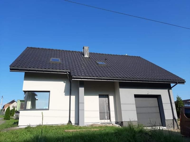 Nowo wybudowany dom jednorodzinny z szarą elewacją, ciemną dachówką i garażem, widoczny z niskiej perspektywy na tle błękitnego nieba.