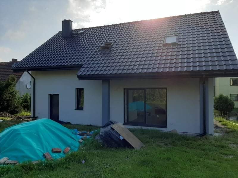 Dom jednorodzinny z szarą elewacją i ciemną dachówką, widoczne przygotowania do prac wykończeniowych, przykryte materiały budowlane przed budynkiem.