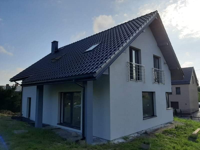 Świeżo otynkowany dom jednorodzinny z ciemną dachówką, widoczne balkony z metalowymi barierkami, w tle drugi budynek.