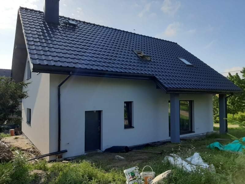 Nowoczesny dom jednorodzinny z szarą elewacją i ciemną dachówką, widoczne ślady prac wykończeniowych na trawniku, w tym wiaderka po farbie i folia zabezpieczająca.