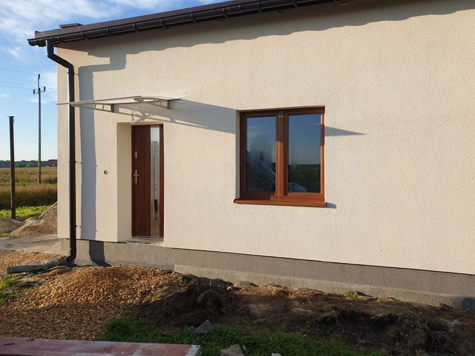 Nowo wykończona elewacja domu jednorodzinnego z drewnianą stolarką okienną i zadaszeniem nad wejściem, widoczne świeże obsypanie ziemią wokół fundamentów.